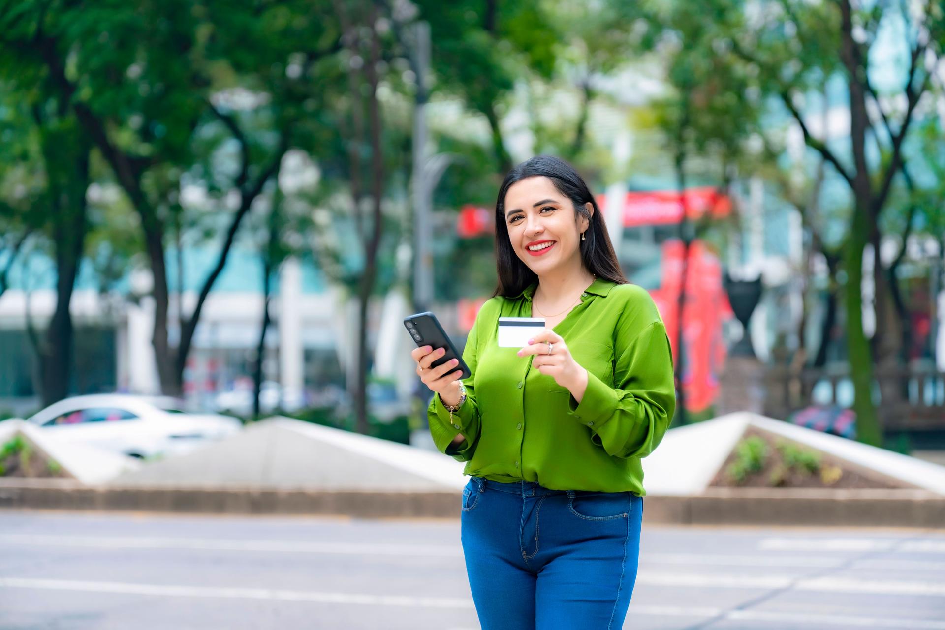 Young Latina woman in green blouse holding credit card and smartphone, smiling outside corporate buildings on Paseo de la Reforma in Mexico City